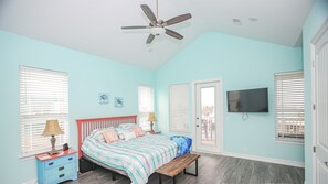 A bedroom with a bed, nightstand, lamp, bench, ceiling fan, TV, and windows. - Kalm by the Beach - Ocean View - Sleeps 14 (Surfside Beach)