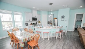 A modern kitchen with white cabinets, a central island, and a dining area with wooden chairs and a striped tablecloth. - Kalm by the Beach - Ocean View - Sleeps 14 (Surfside Beach)