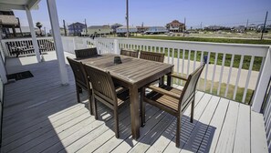 Una terraza de madera con una mesa y sillas, baranda blanca, y vistas a casas y una carretera al fondo.