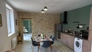 A modern kitchen with a stone wall, a dining area with a table and chairs, a washing machine, and a view of a green outdoor area. - La Forge, house for 6 in the heart of Barfleur, with private parking lot (Barfleur)