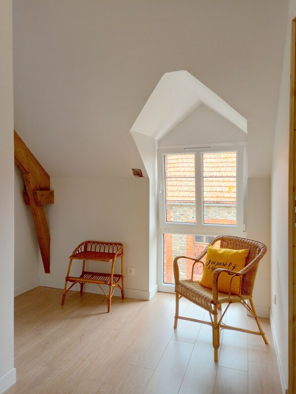 A room with a sloped ceiling, a wicker chair with a cushion, a small wooden table, and a window with a view of a brick building.