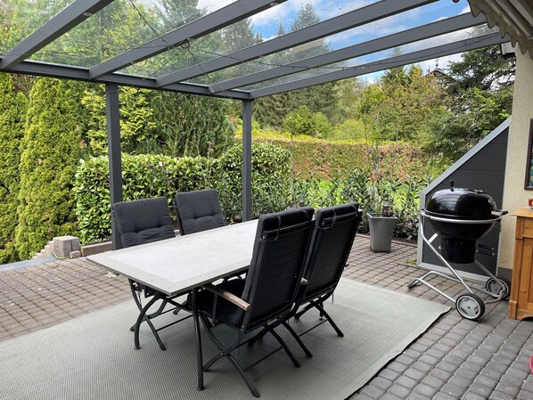 A covered patio with a table and chairs, a grill, and a view of a garden.