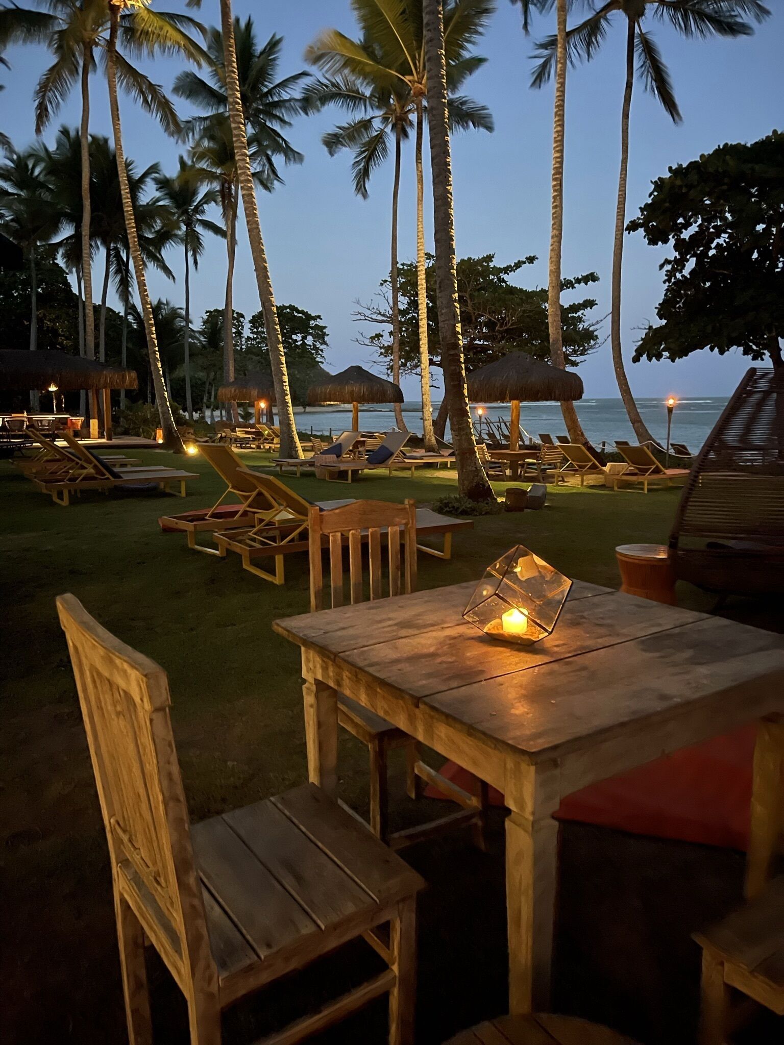 A wooden table with chairs and a lantern, surrounded by palm trees and beach views.
