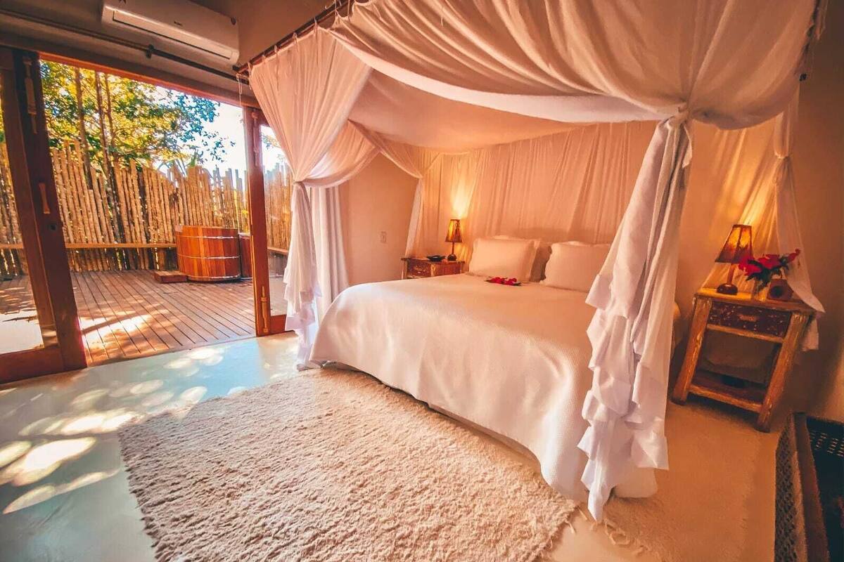 Een kamer met een hemelbed, houten vloeren, uitzicht op bamboe en een klein nachtkastje met een lamp.