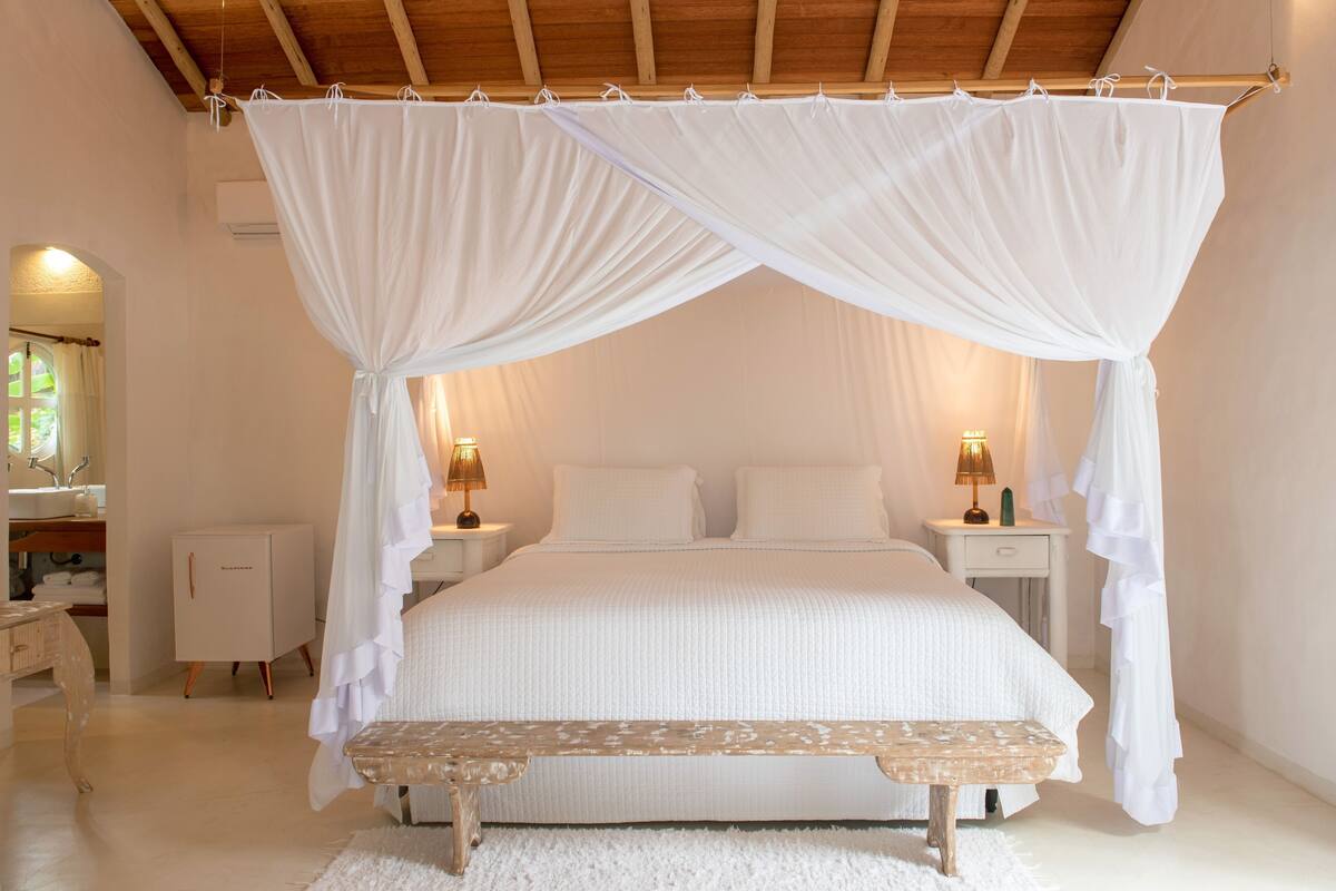 Een slaapkamer met een hemelbed, nachtkastjes met lampen, een bankje en een garderobe.