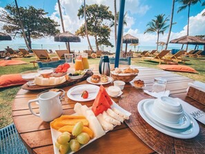 Een ontbijtbuffet op een houten tafel met uitzicht op tropische palmbomen en de oceaan.