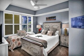 A bedroom with a bed, nightstands, a ceiling fan, a window with shutters, and a wall-mounted sign.