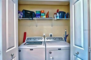 A laundry room with two front-loading washing machines, a shelf above with cleaning supplies, and a door on the right.