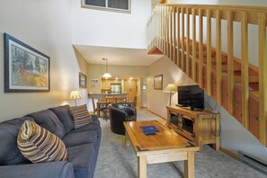 Relax in this inviting living area with a sofa, coffee table, and TV, plus a staircase leading to an upper level and a kitchen area.