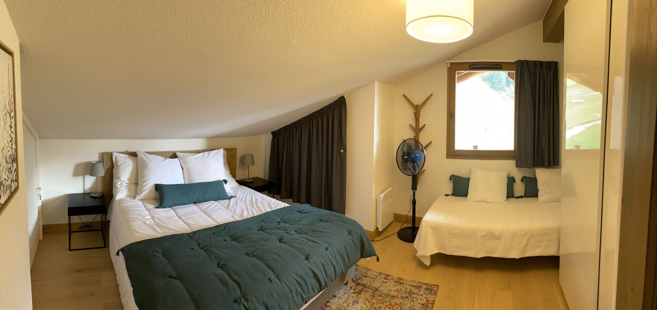 A bedroom with a large bed, a bedside table, a standing fan, and a window with curtains.