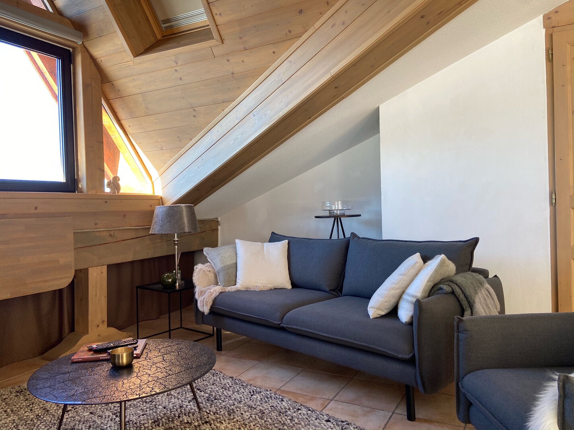 Une pièce confortable de style loft, avec un plafond en bois, un canapé gris agrémenté de coussins blancs et gris, une petite table ronde avec une lampe, et une vue sur l’extérieur par une fenêtre.