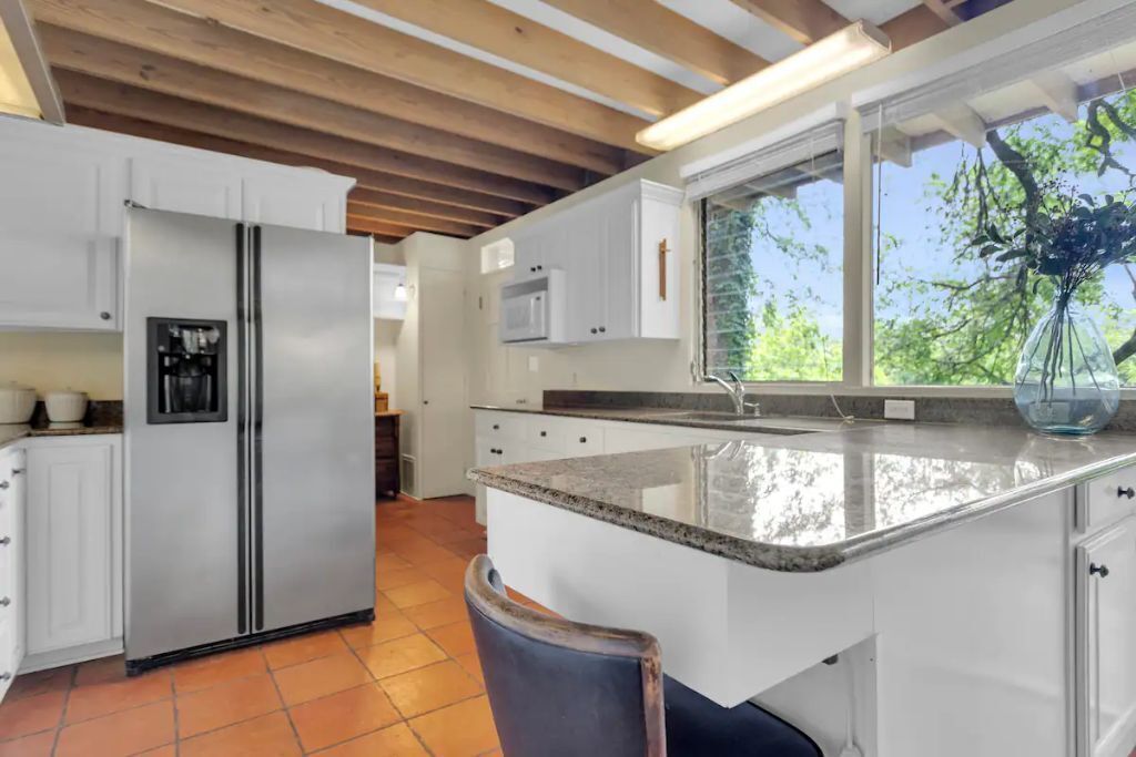 A modern kitchen with white cabinetry, a stainless steel refrigerator, a granite countertop, and a window with a view of greenery.