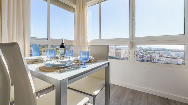 Zona pranzo con tavolo apparecchiato per due persone, una bottiglia di vino e vista sulla città attraverso ampie finestre.