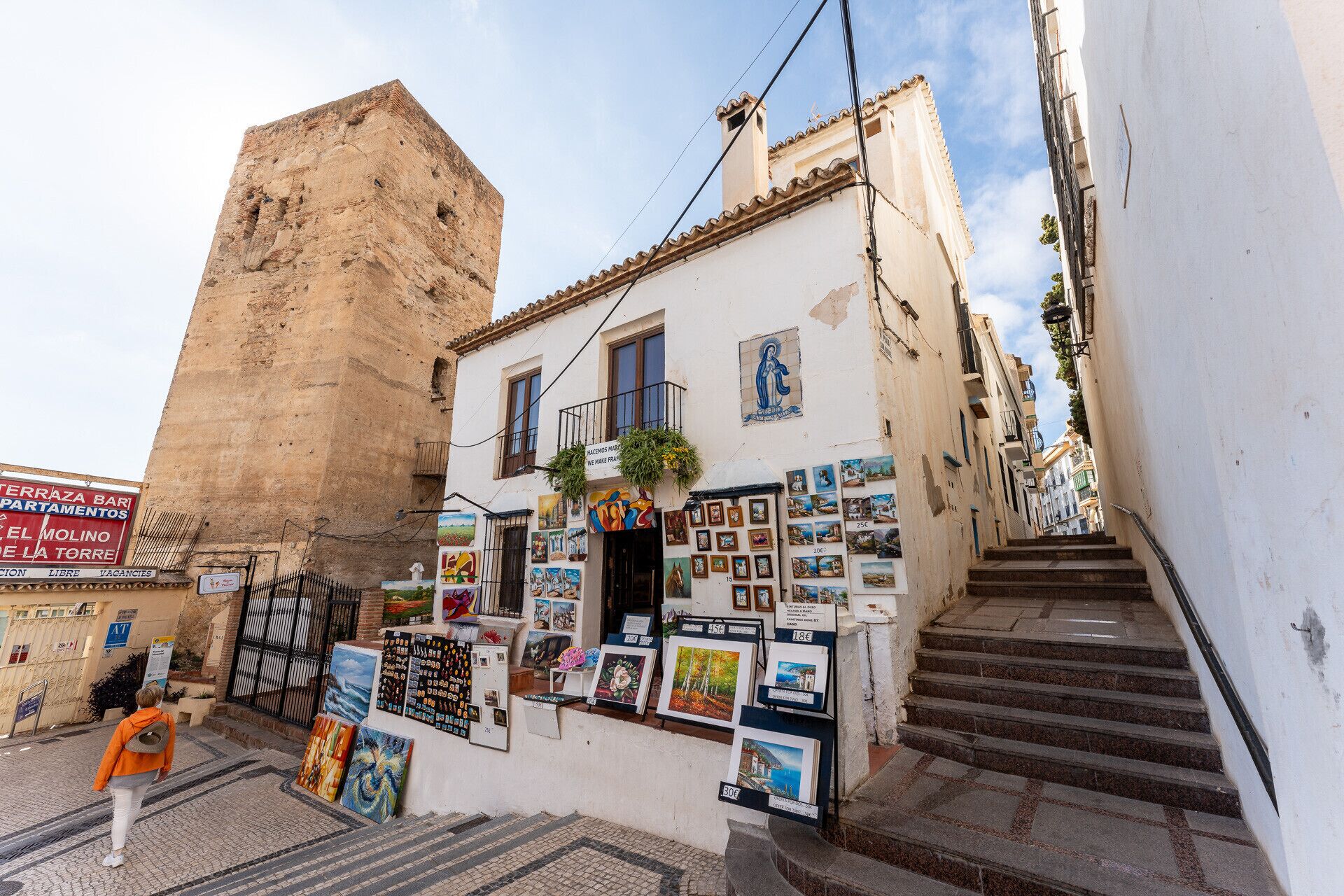 A narrow street with a building displaying various artworks and a staircase leading to a higher level.