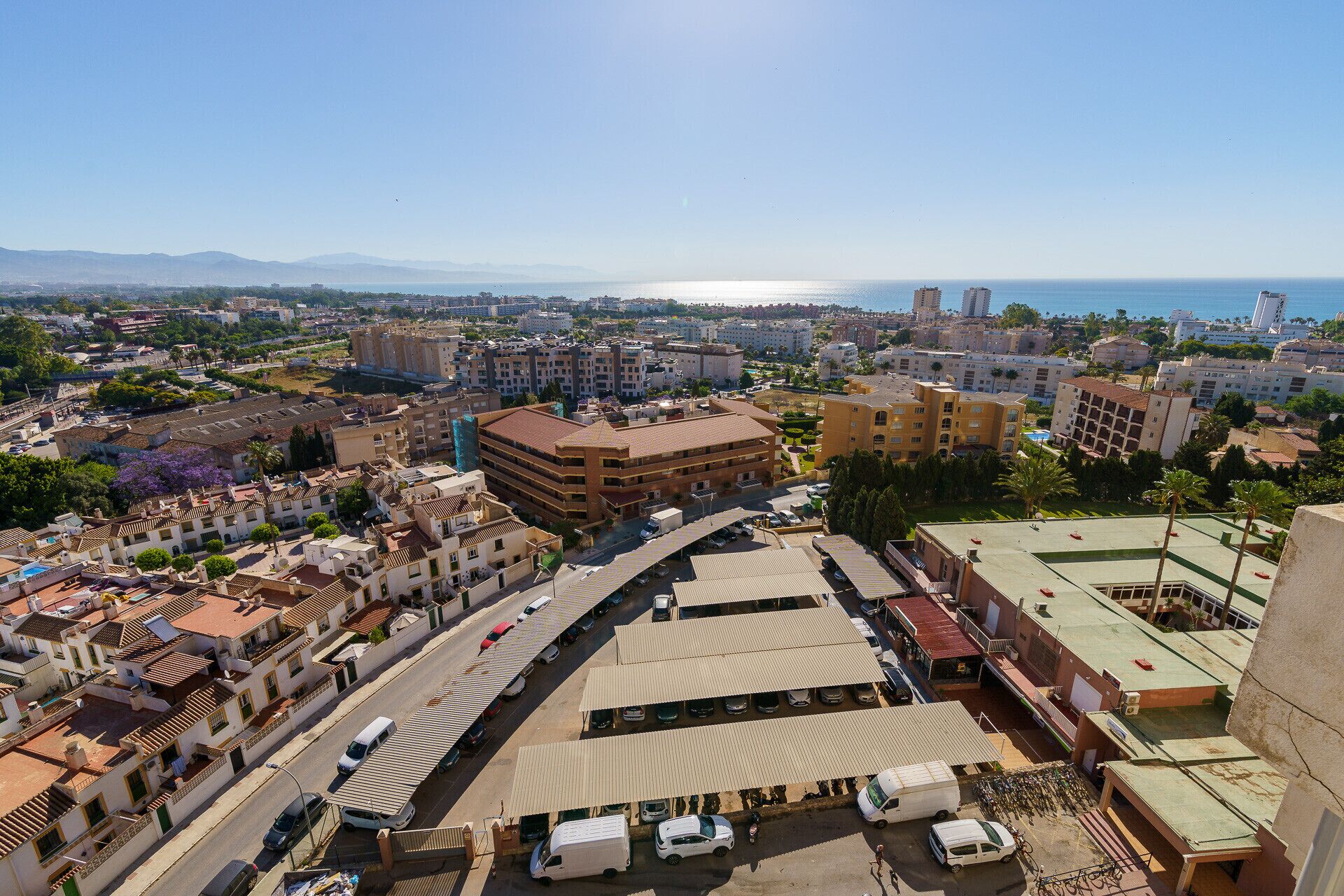 Vue aérienne d’une ville côtière, avec un mélange de bâtiments résidentiels et commerciaux, un imposant bâtiment circulaire, et une vue dégagée sur la mer et les montagnes au loin.