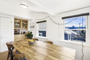 A dining area with a wooden table, chairs, a bowl of fruit, a cabinet with kitchen items, and a window with a view of buildings.