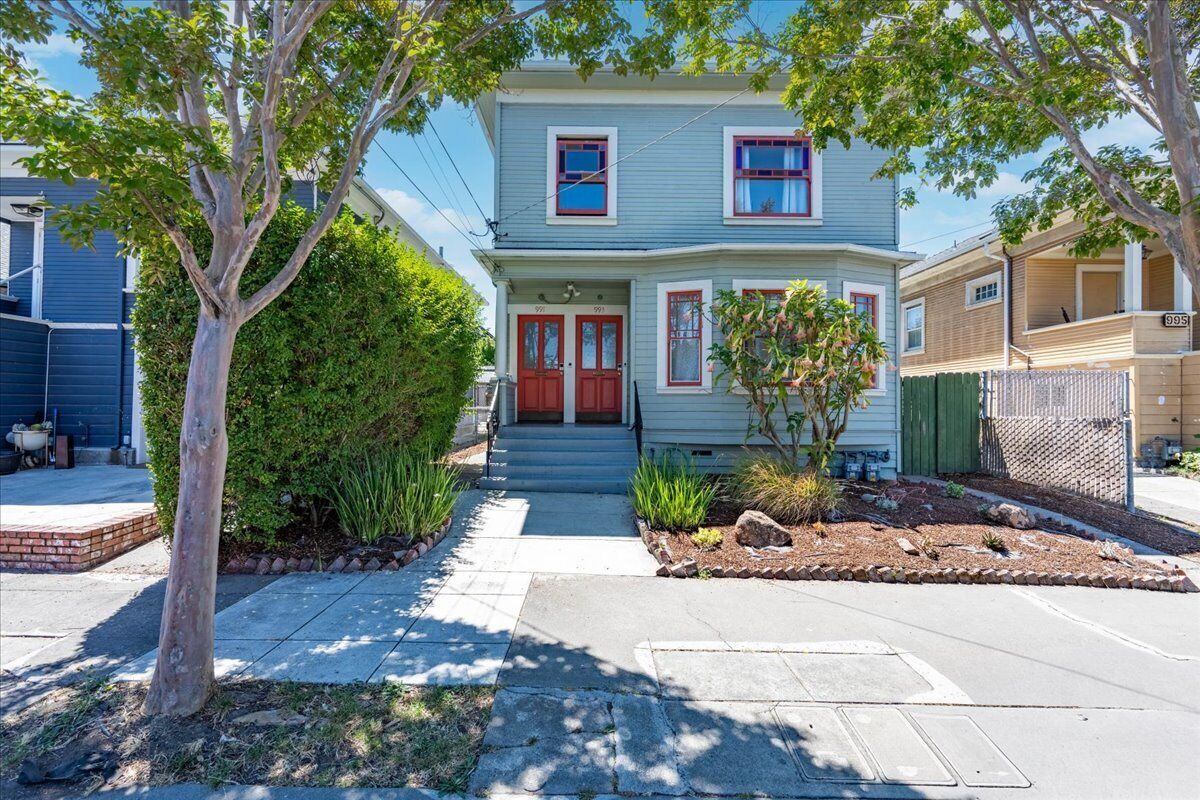A two-story house with a red door, a small front yard with plants, and a sidewalk leading to the entrance.