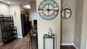 A spacious living room with a large wooden floor, a dining area with a black chair and table, a wall clock, and a small side table with decorative items. - Gulfcrest Townhomes Unit 3 (Destin)
