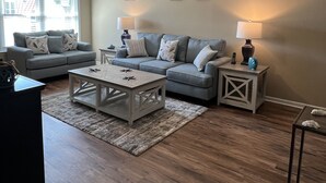 A living room with a high ceiling, a large window, a grey sofa set, a wooden coffee table, and two side tables with lamps. - Gulfcrest Townhomes Unit 3 (Destin)