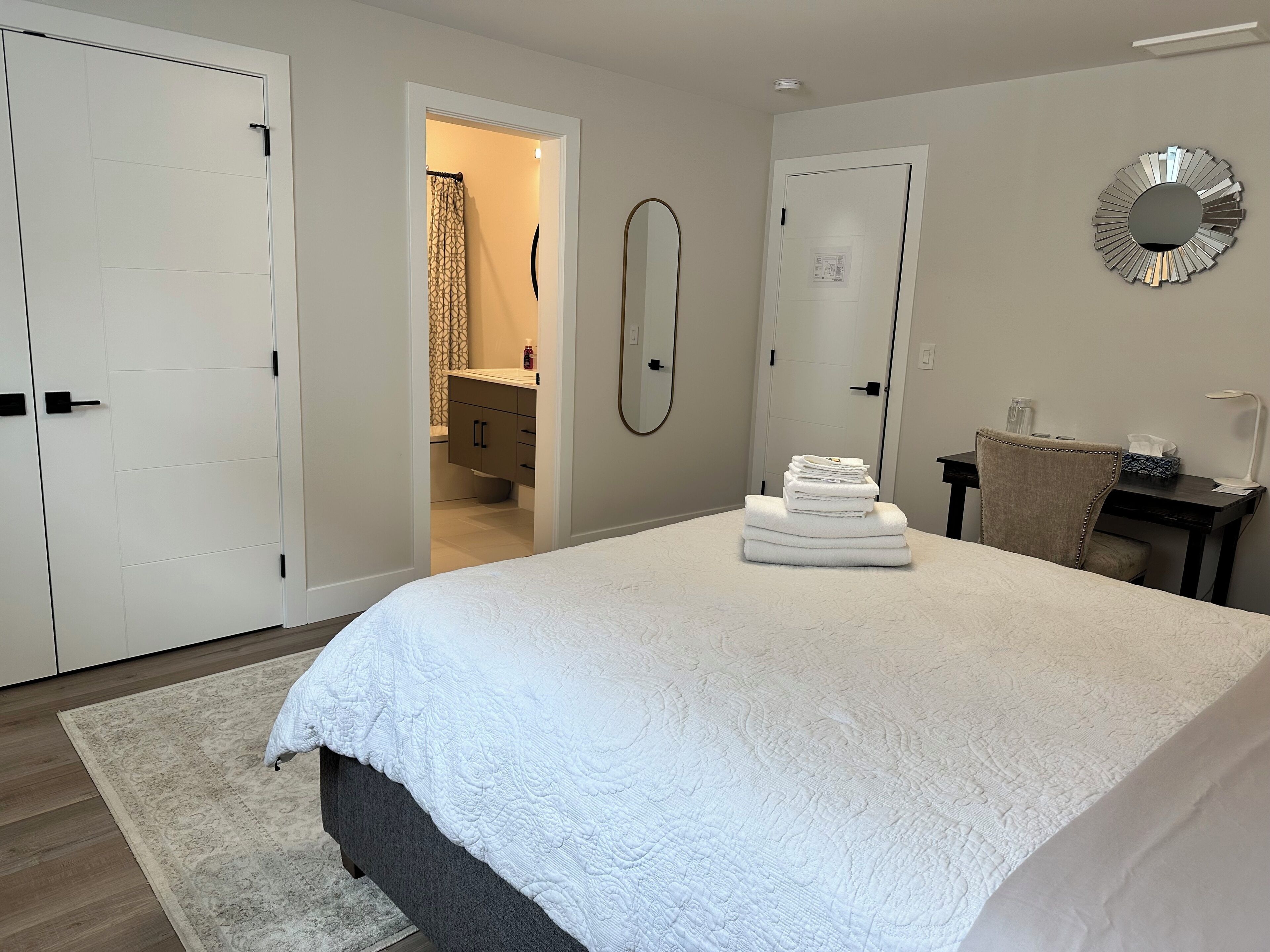 A bedroom with a large bed, a bedside table with a lamp, a chair, and a desk with a mirror.