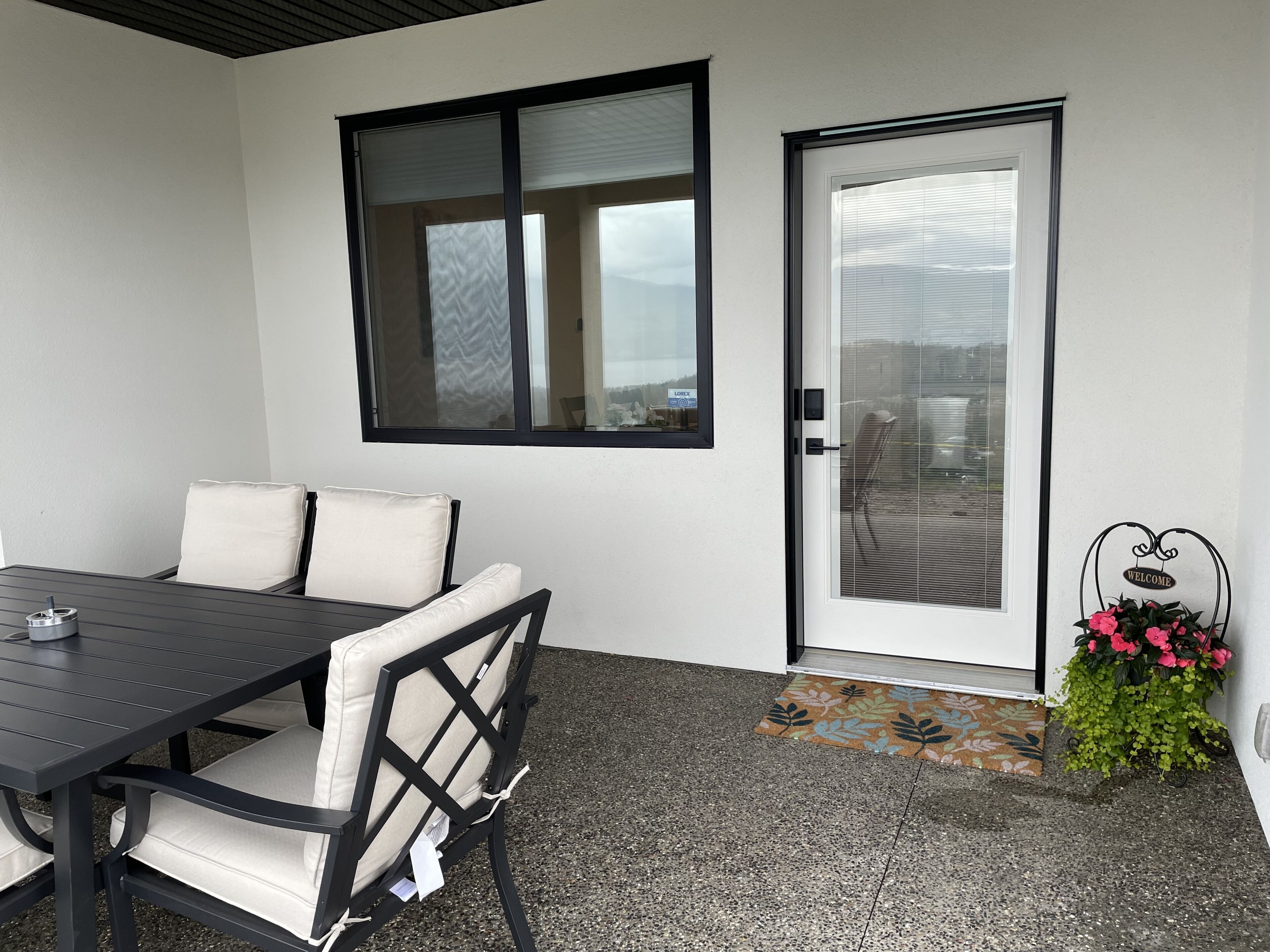 Un área de estar moderna al aire libre, con una mesa, sillas, un gran ventanal corredero y un letrero de bienvenida con flores.
