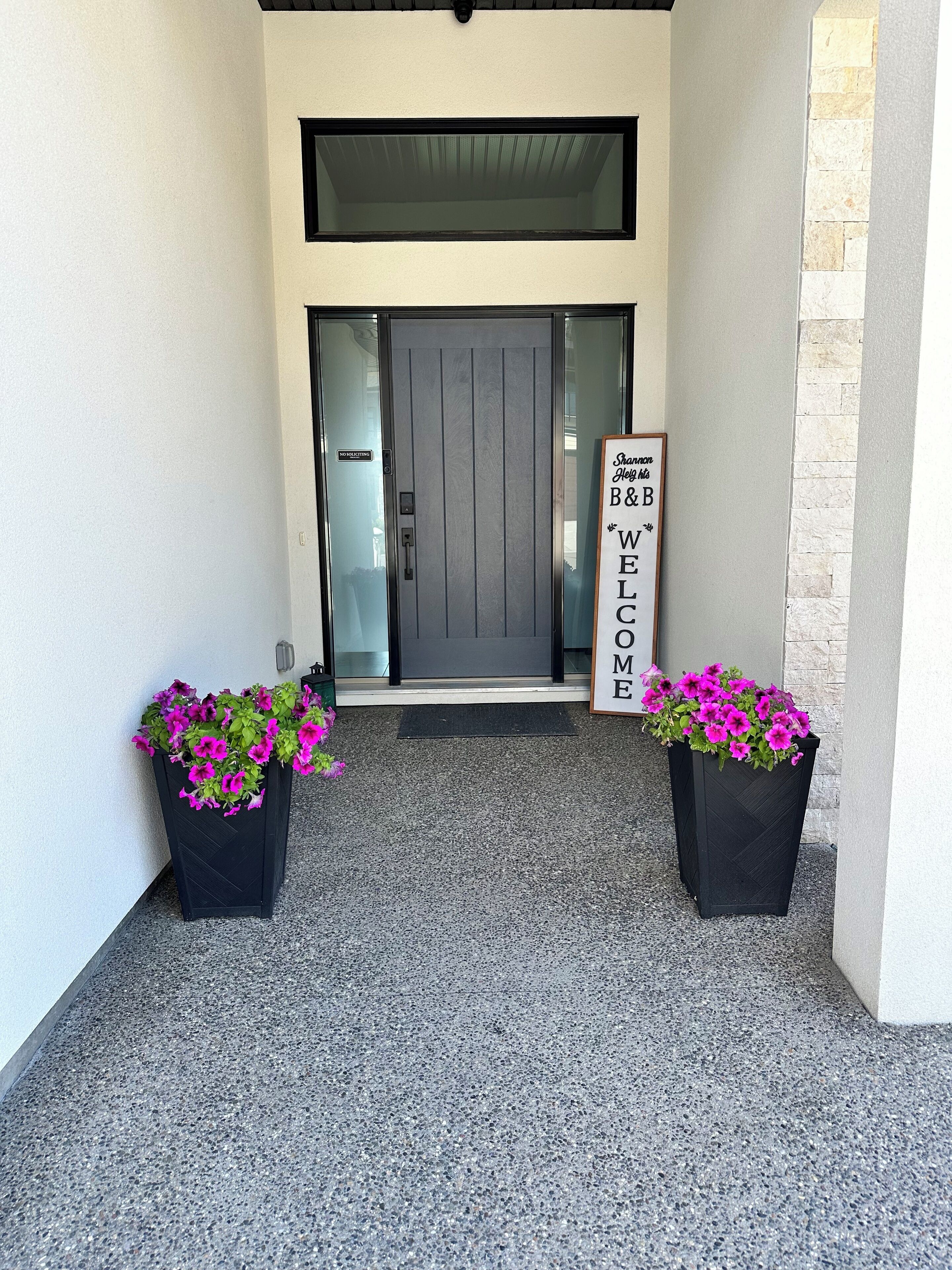A modern entrance with a glass door, two black planters with pink flowers, and a sign with the text "Welcome Home."