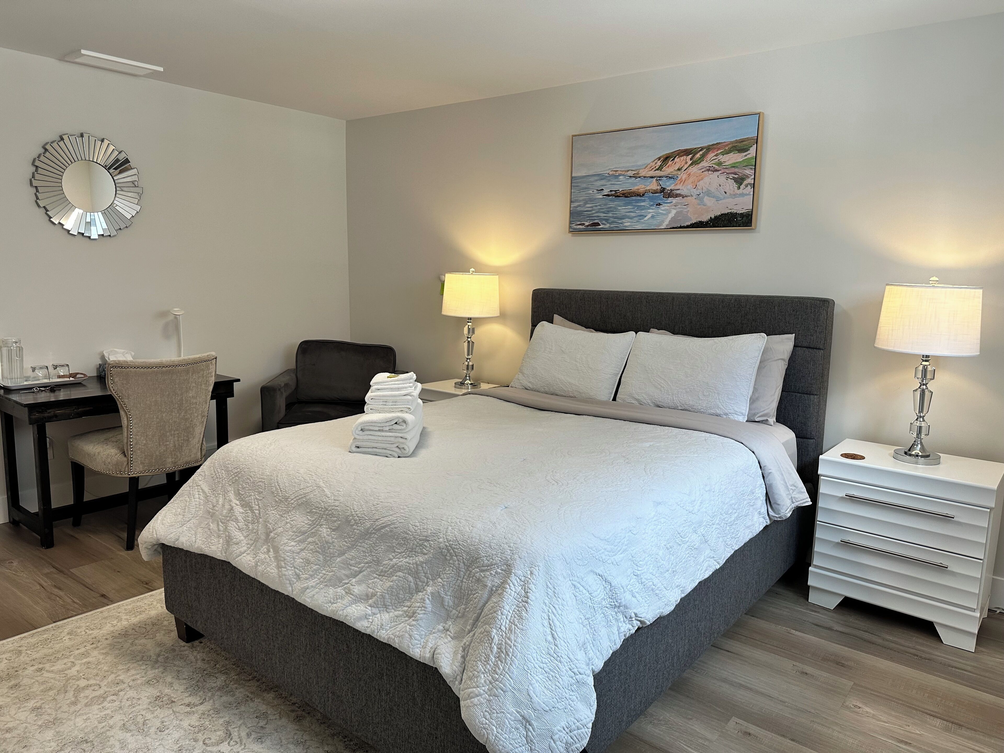 A neatly arranged hotel room with a large bed, a desk with a chair, a nightstand with a lamp, and a painting on the wall.