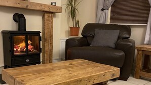 A living room with a wooden coffee table, a black sofa, a fireplace, a potted plant, and a window with curtains. - Riverbank Cottage Lake District, Double Balcony (Egremont)