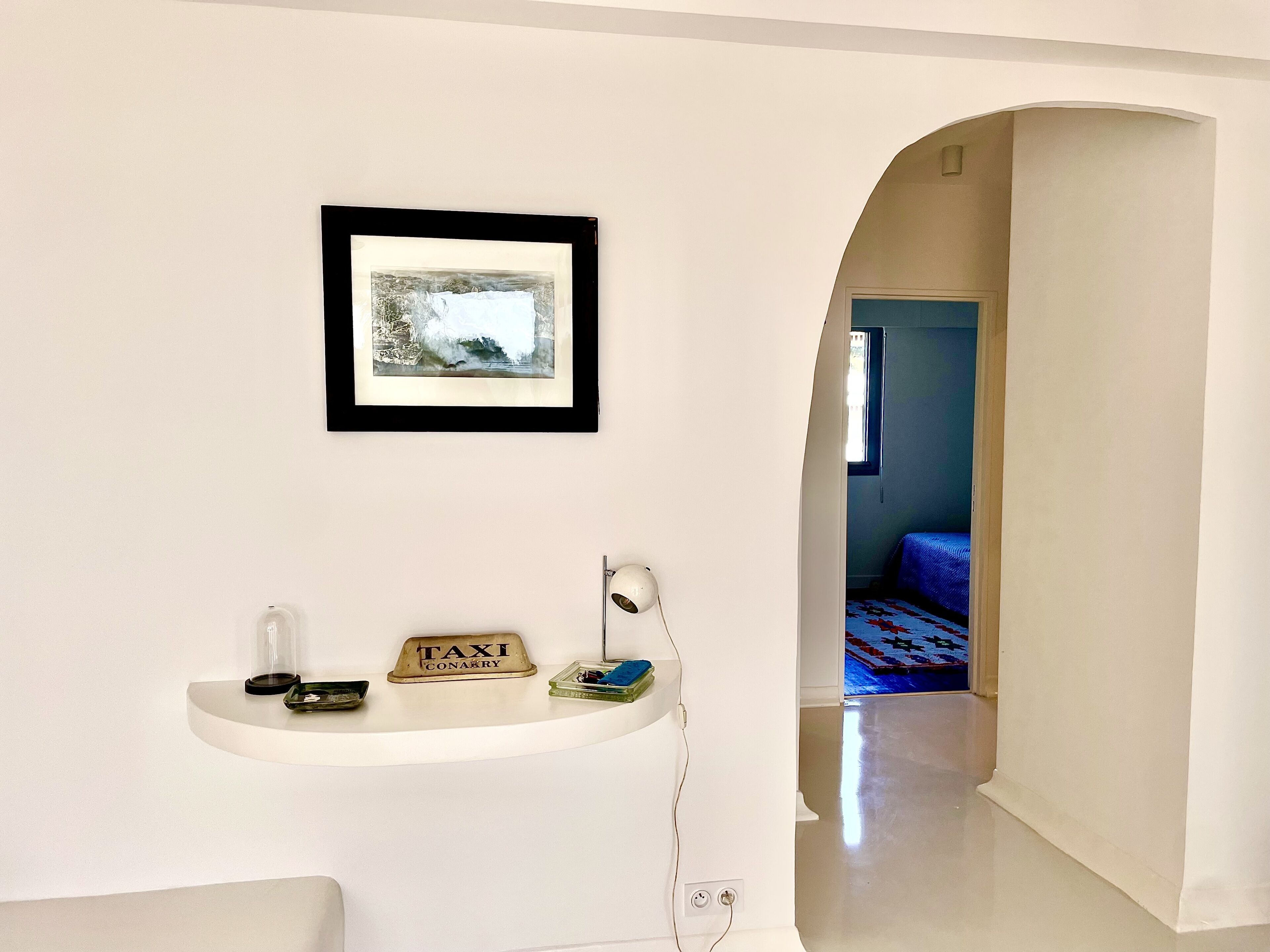 Une pièce avec un tableau encadré au mur, une petite étagère sur laquelle se trouve un panneau indiquant « taxi », une lampe et une prise de courant.