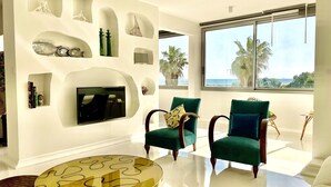 Ein modernes Wohnzimmer mit Kamin, zwei grüne Sessel, einem runden Glastisch und einem großen Fenster mit Blick auf Palmen und das Meer.