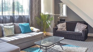 A living room with a grey sofa, a glass coffee table, and a grey armchair. There is a potted plant and a lamp on a side table. - Huddersfield House, Wood View (Huddersfield)