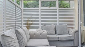 A conservatory with a cushioned seating area, large windows, and a view of greenery. - Huddersfield House, Wood View (Huddersfield)