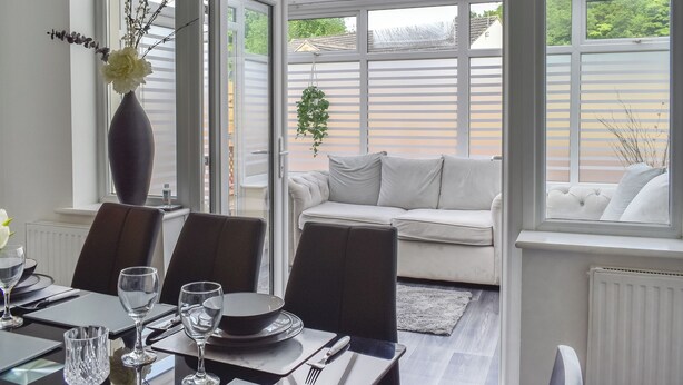 A dining area with a table set for four, a sofa, and a view of a garden through large windows. - Huddersfield House, Wood View (Huddersfield)
