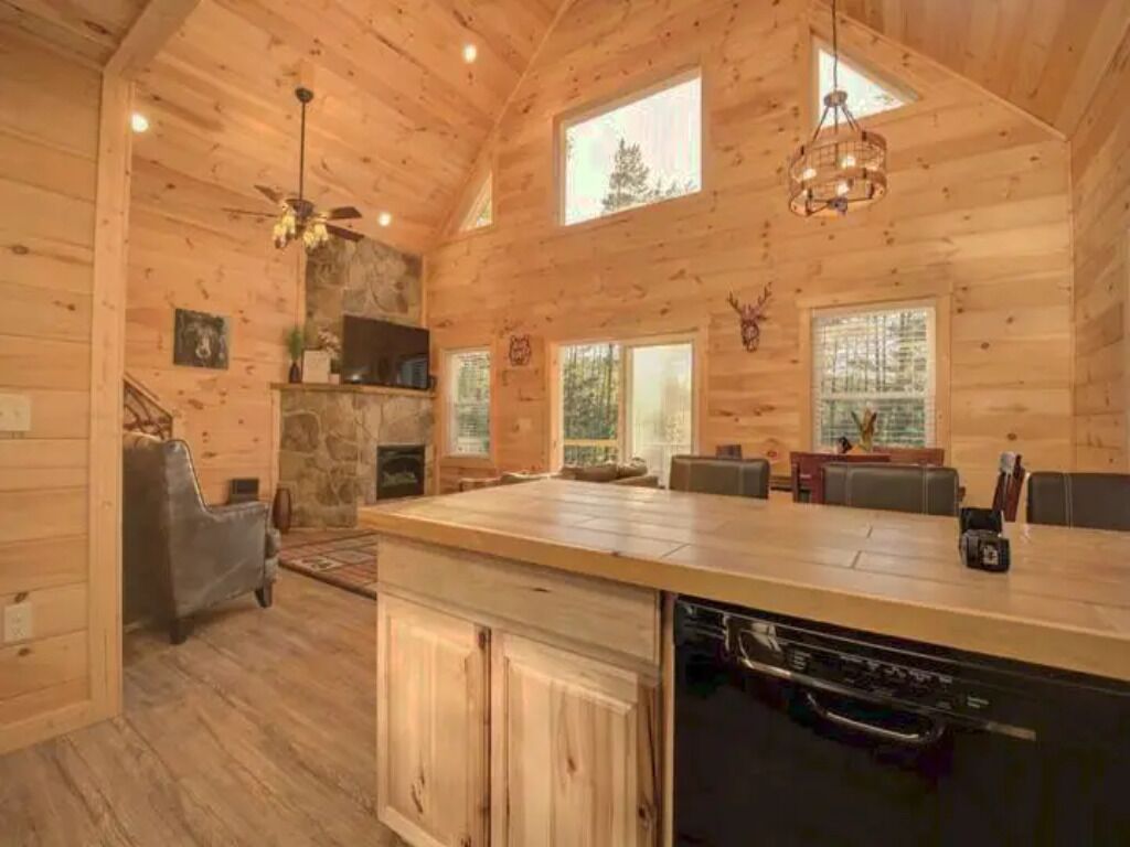 Un intérieur de chalet en bois spacieux, doté d’un grand îlot de cuisine, d’une cheminée en pierre et de nombreuses fenêtres offrant une vue sur la forêt.
