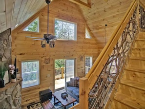 A wooden cabin interior with a stone fireplace, a ceiling fan, a staircase, and a sofa.