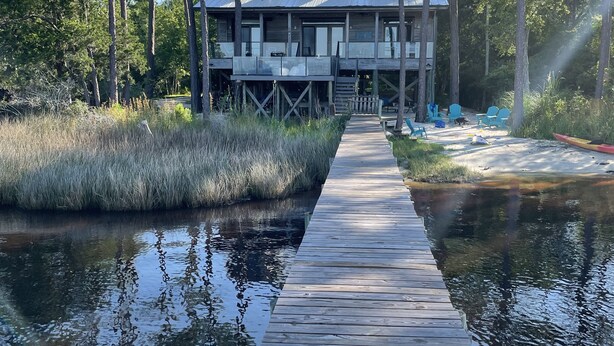 A wooden dock leading to a two-story house with a covered deck area, surrounded by tall trees and a calm body of water. - Sunsets on Water - private beach, pier, boat dock, fishing -12m to Navarre Beach (Navarre)