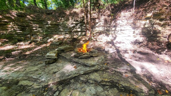 A natural outdoor setting with a fire burning on rocks, surrounded by trees and a stone wall. - ❤️ Tiny House with Creek Access ❤️ (Anderson)