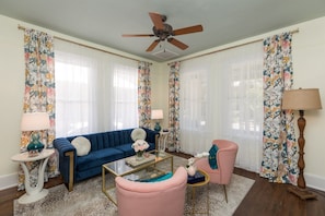 Una sala de estar con un sofá azul, un sillón rosa, una mesa de centro de vidrio y un ventilador de techo.