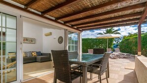 Un patio exterior con mesa de vidrio y sillas de mimbre, un sofá, y una piscina con sillones de descanso y una sombrilla.