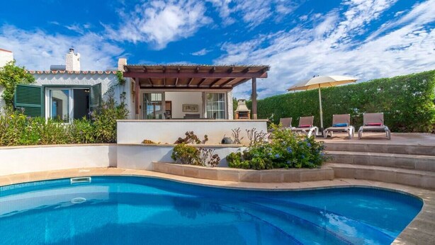 Una piscina con una pérgola y sillones de descanso, rodeada de un jardín ajardinado y un edificio con techo de tejas.