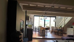 Àmplia zona d'estar amb un gran finestral, zona de dinar amb cadires i taula, i una cuina visible al fons.