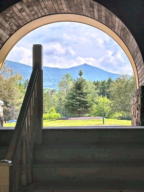 A view through an arched window with a staircase leading to a grassy area, surrounded by trees and mountains.