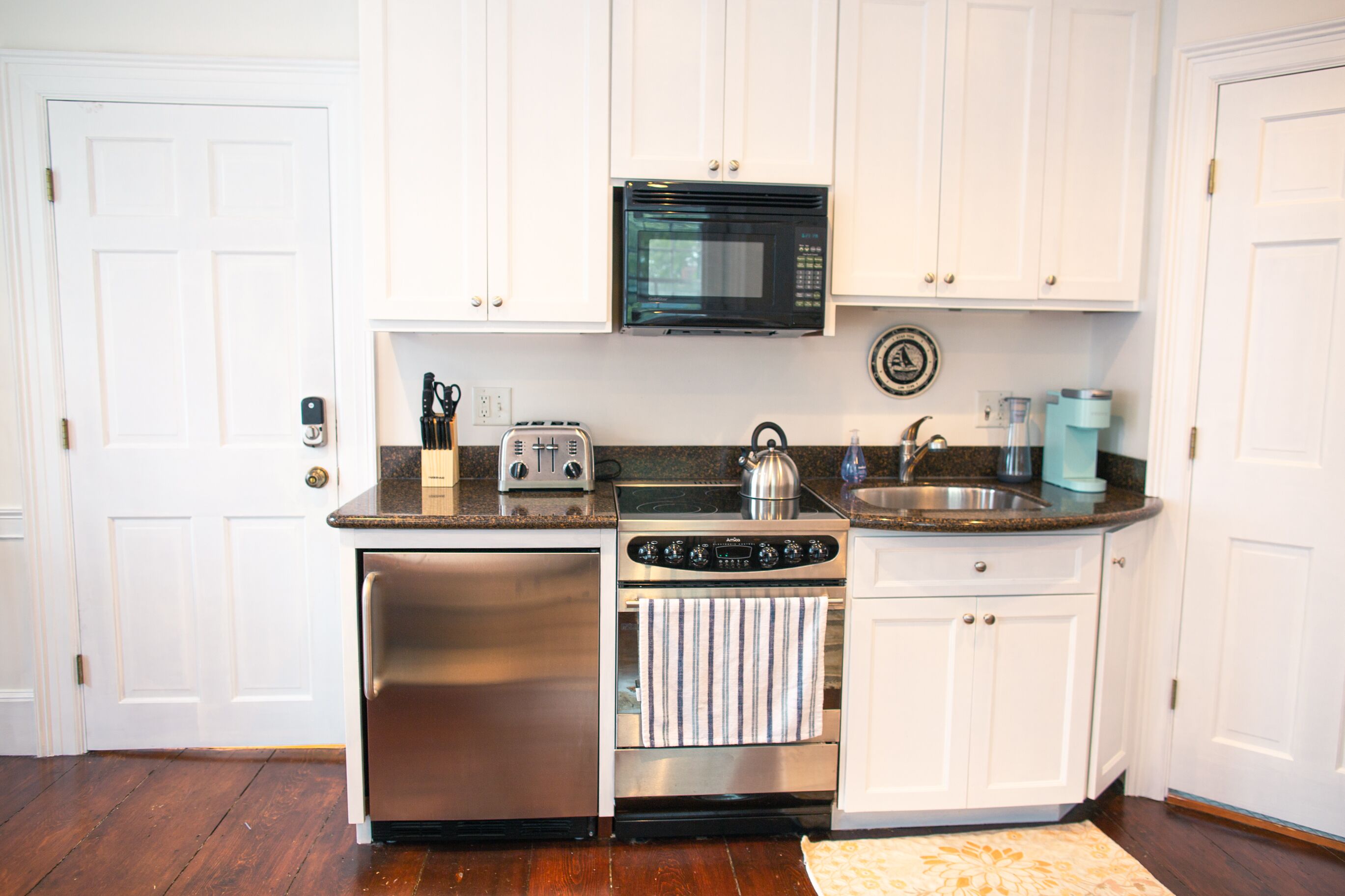 A compact kitchen with white cabinets, a stainless steel refrigerator, a microwave, a toaster, a kettle, and a sink.