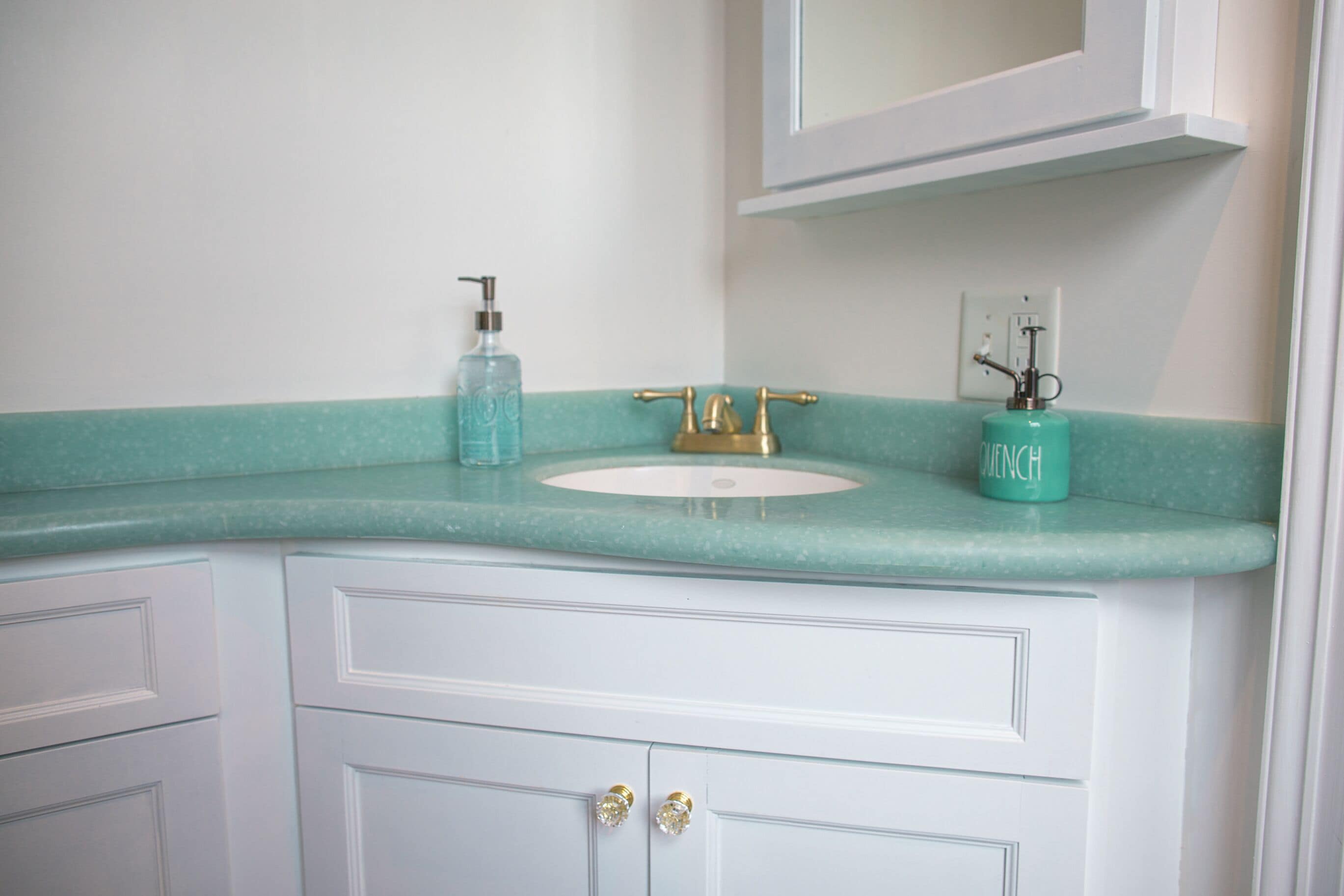 Een badkamer met een wit wastafelmeubel, een turquoise aanrechtblad, een gouden kraan en een zeegroene zeepdispenser.