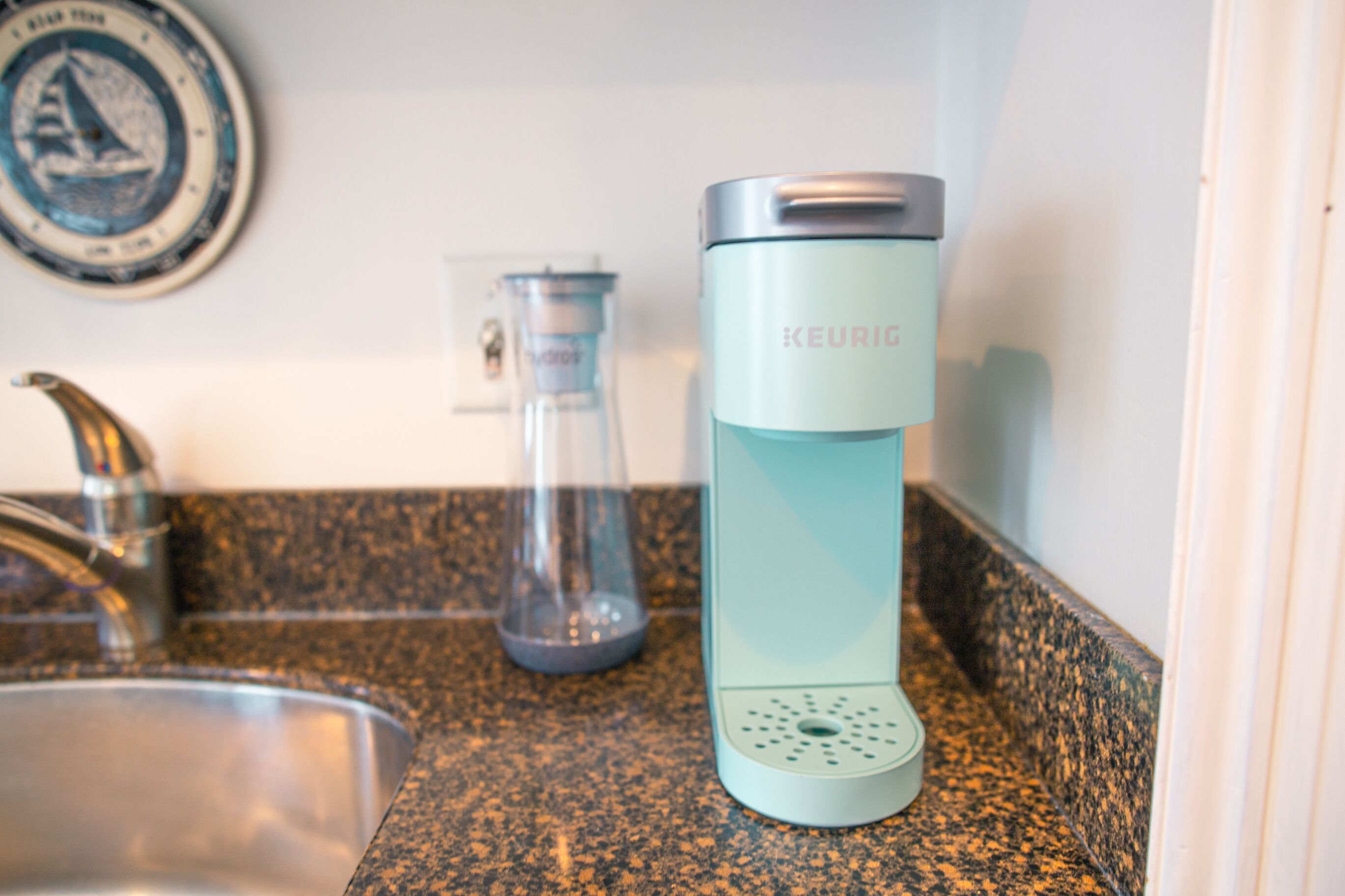 A kitchen counter with a sink, a glass carafe, and a teal-colored electric kettle.