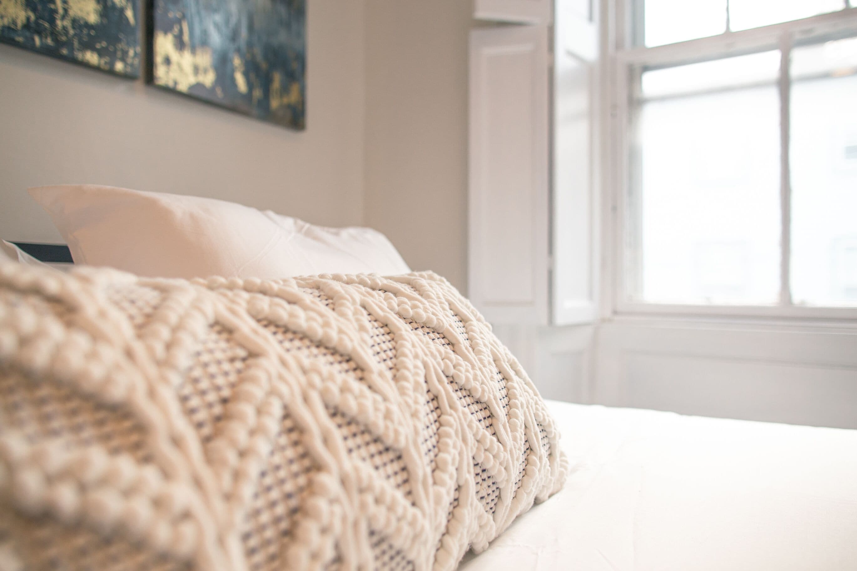 Una cama con una manta tejida y almohadas en una habitación con ventana y cuadros en la pared.