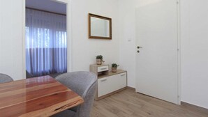 Una habitación con una mesa de comedor de madera, sillas tapizadas grises, un mueble blanco pequeño con dos plantas en macetas, un espejo en la pared y una puerta blanca.