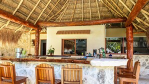 A bar area with wooden chairs and a stone counter, a thatched roof, and a view of the outdoors. - Villa Marcela (Chetumal)