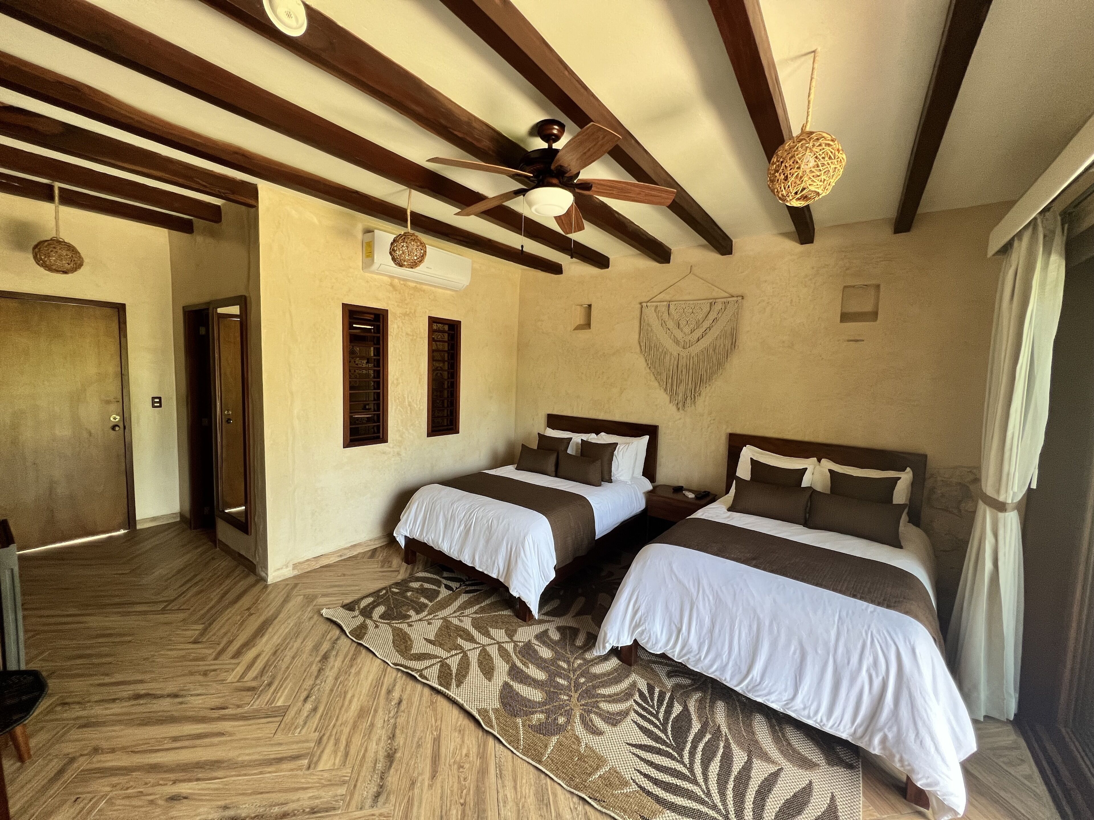 Habitación de hotel con dos camas, pisos de madera, un ventilador de techo y un tapiz de pared tejido.