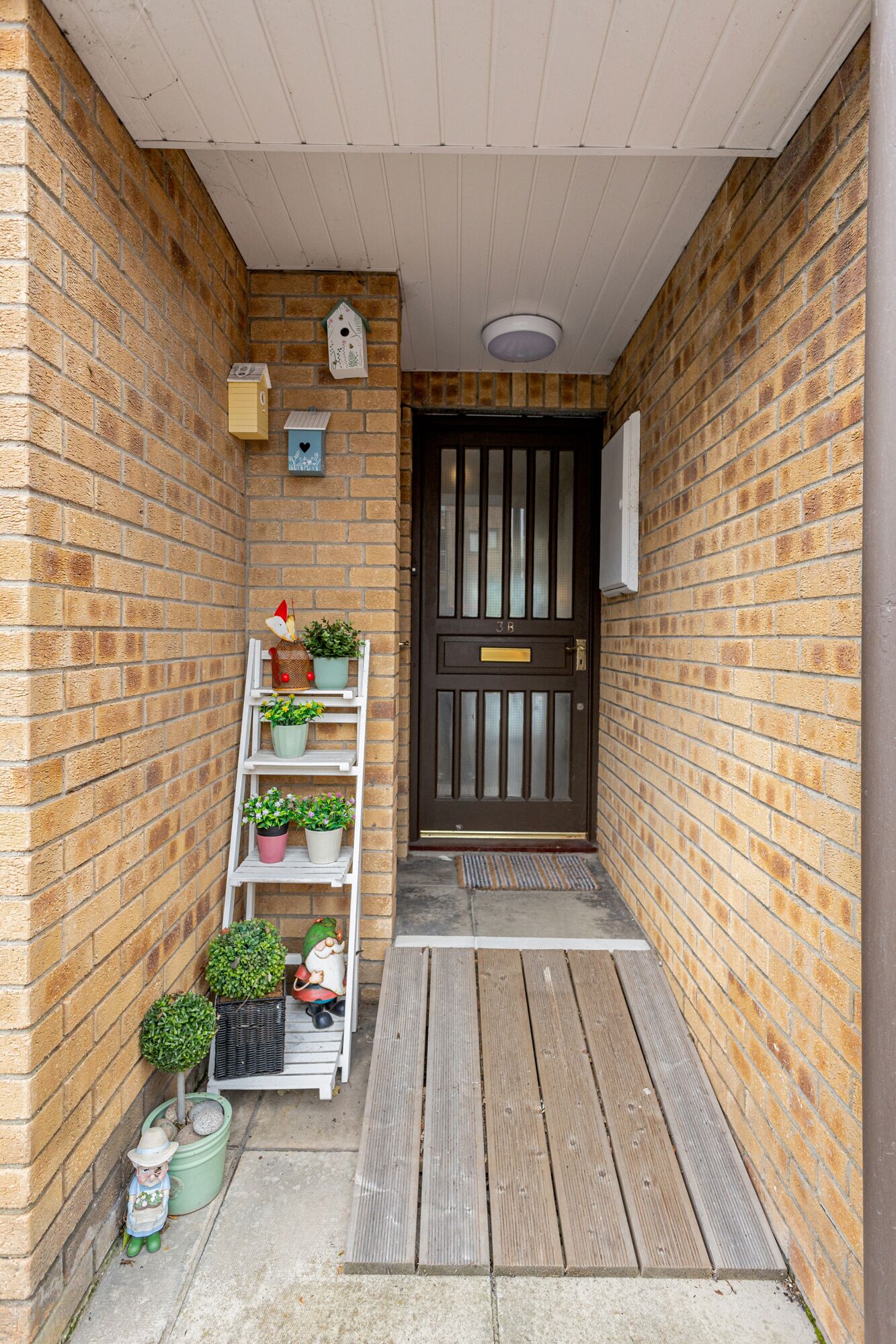 A narrow brick corridor with a wooden bench, a door with a brass plate, and a white shelf with potted plants.